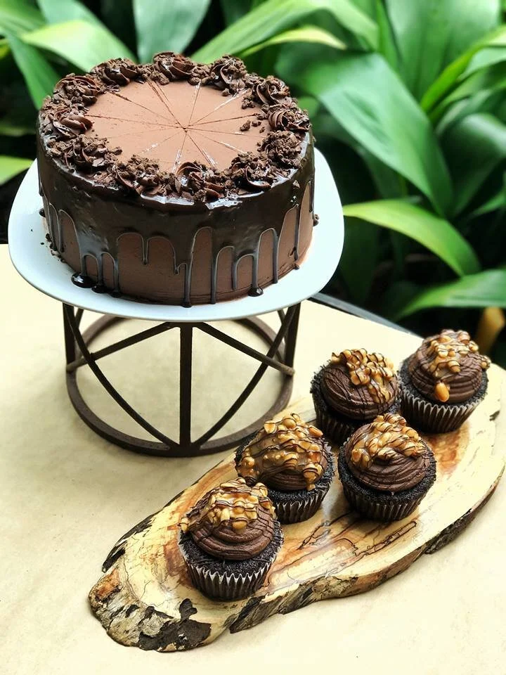 A chocolate cake with dark ganache running down the sides, on a stand beside frosted cupcakes