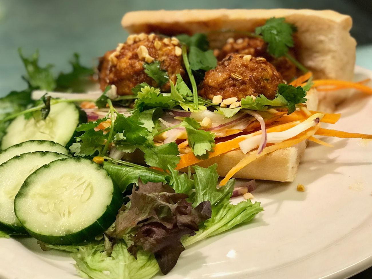 A fried chicken sandwich with pickled carrot and coriander, beside cucumber and salad leaves